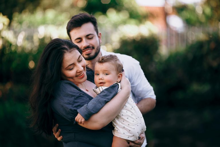 Un plano medio captura a una mujer sonriente sosteniendo a un niño pequeño, con un hombre sonriente de pie muy cerca detrás de ellos al aire libre. La mujer, que tiene el pelo largo y oscuro y lleva una blusa azul oscura, mira con cariño al niño. El hombre, con camisa blanca, está de pie con los ojos cerrados y una expresión amable. El niño, vestido con un peto claro estampado, mira ligeramente fuera de cámara. El fondo consiste en un follaje verde suave y desenfocado.