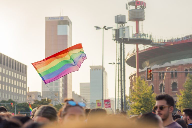 Bandera arcoíris ondeando durante una manifestación del Orgullo LGTBIQ+ en una ciudad, con multitud de personas y edificios urbanos al fondo.