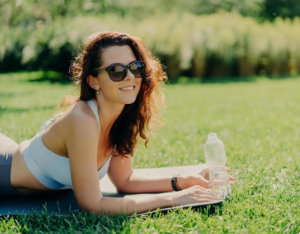 Foto de una mujer morena alegre y relajada posa sobre una colchoneta de fitness, usa gafas de sol de moda y ropa deportiva, bebe agua y siente sed después de los ejercicios deportivos, tumbada sobre césped verde y disfruta de un buen día soleado.